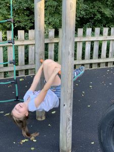 Olivia hangs upside down from some play equipment. Her knees are tucked over a bar which she also hangs on to with her hands
